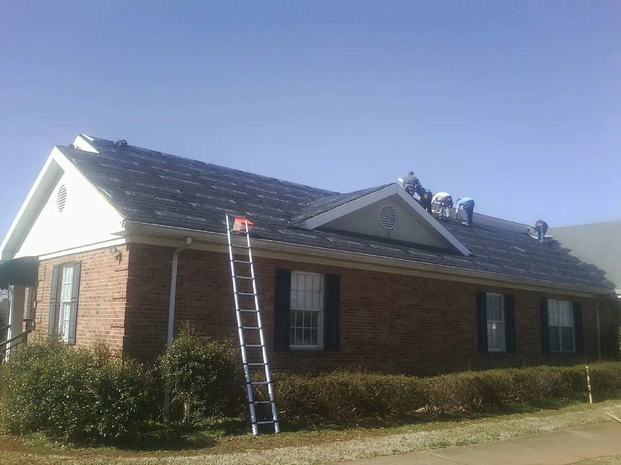 Crew tearing shingles off a brick ranch roof with a ladder set — Metal Roof Repair work by our crew in Alva